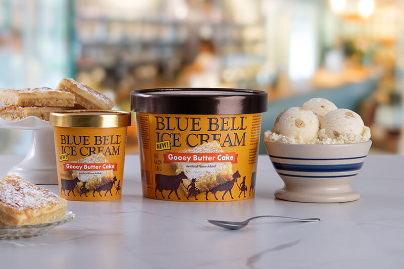 Blue Bell Gooey Butter Cake Ice Cream in half gallon and pint with a bowl sitting on a bakery counter with gooney butter cake pieces