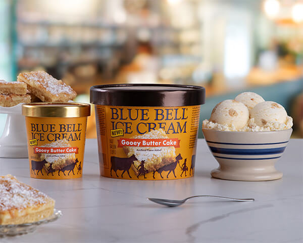 Blue Bell Gooey Butter Cake Ice Cream in half gallon and pint with a bowl sitting on a bakery counter
