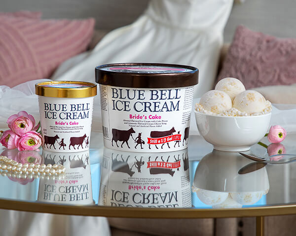 Blue Bell Bride's Cake Ice Cream in half gallon and pint. Ice cream in bowl on glass table with wedding dress, pink flowers, veil, and pearl necklace.